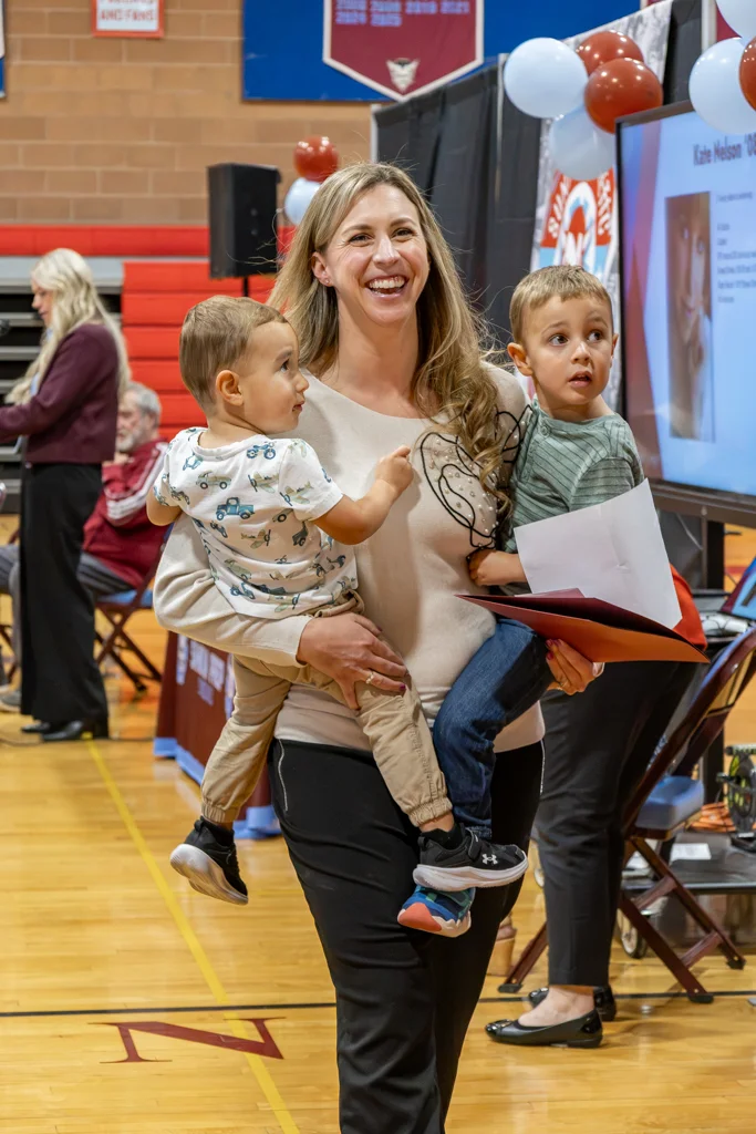 Woman walks across stage holding two toddlers.